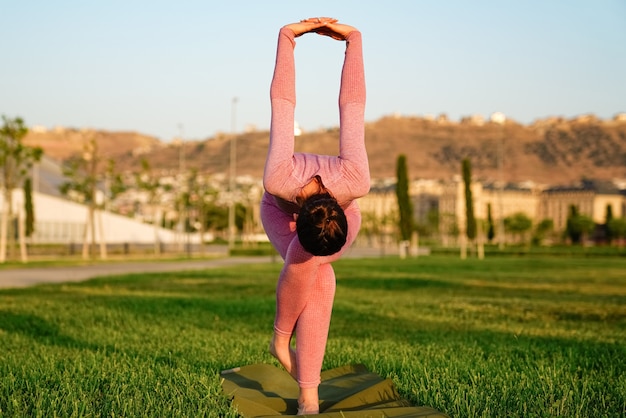 Mujer joven en camisa rosa y pantalones en la hierba dentro del parque verde meditando y haciendo yoga en diferentes poses