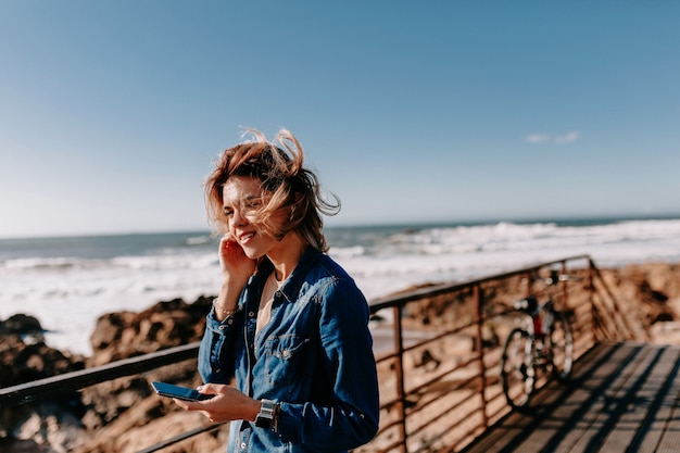 Mujer joven con camisa vaquera está hablando por teléfono posando en la playa