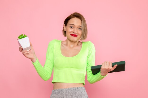 Mujer joven en camisa verde y falda gris sonriendo sosteniendo la planta y el cuaderno