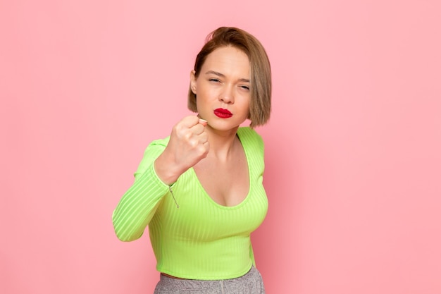 Mujer joven con camisa verde y pantalón gris posando con expresión amenazante