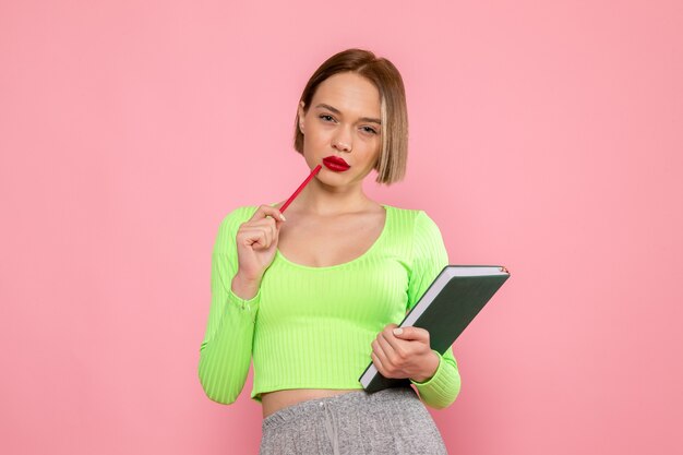 Mujer joven en camisa verde y pantalón gris posando con lápiz rojo y cuaderno