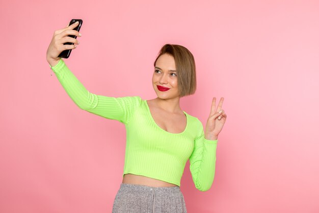 Mujer joven en camisa verde y pantalón gris posando y tomando un selfie