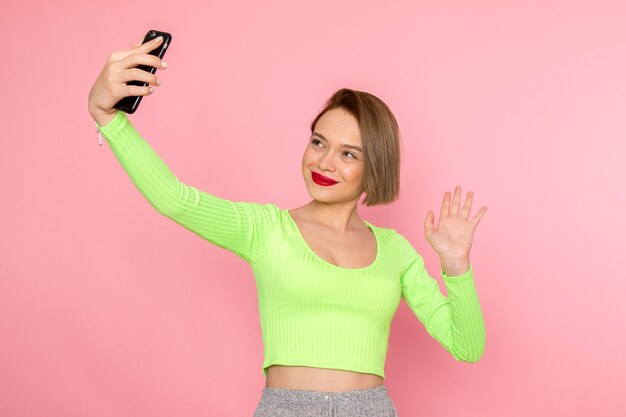 Mujer joven en camisa verde y pantalón gris sonriendo y tomando un selfie