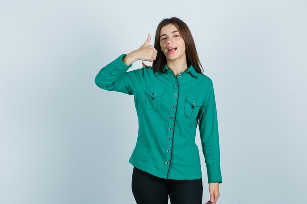 Mujer joven con camisa verde, pantalones mostrando el pulgar hacia arriba mientras parpadea y parece confiado, vista frontal.