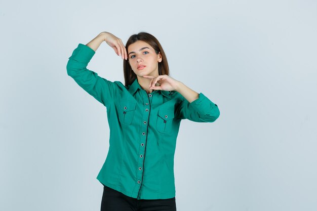 Mujer joven en camisa verde posando con las manos alrededor de la cabeza y luciendo delicada, vista frontal.