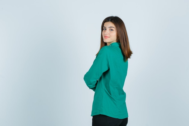 Mujer joven en camisa verde sosteniendo los brazos cruzados mientras mira hacia atrás y mira alegre, vista posterior.