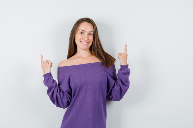 Mujer joven en camisa violeta apuntando hacia arriba y mirando alegre, vista frontal.