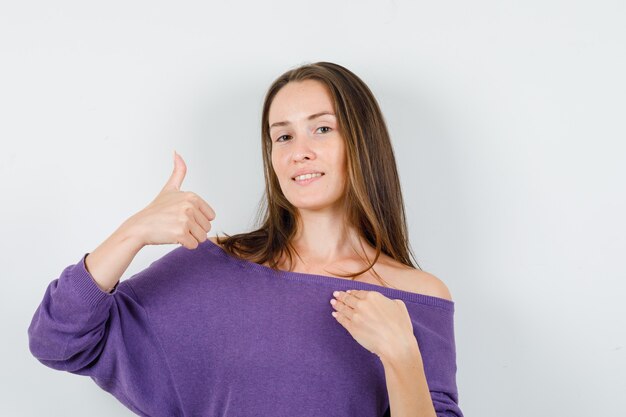 Mujer joven con camisa violeta apuntando a sí misma y mostrando el pulgar hacia arriba, vista frontal.
