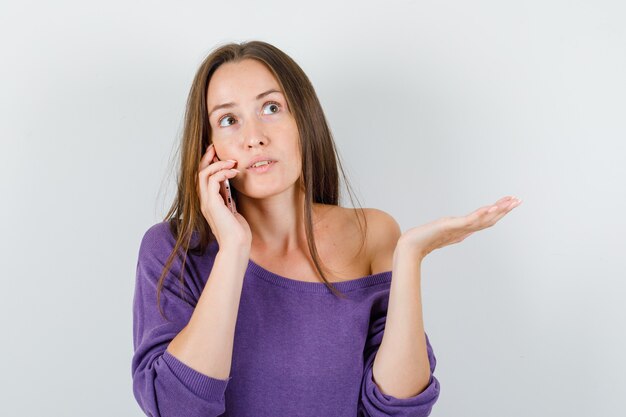 Mujer joven en camisa violeta hablando por teléfono móvil y mirando confundido, vista frontal.