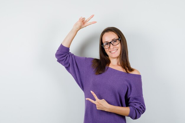 Mujer joven con camisa violeta mostrando el signo de la victoria y con suerte, vista frontal.