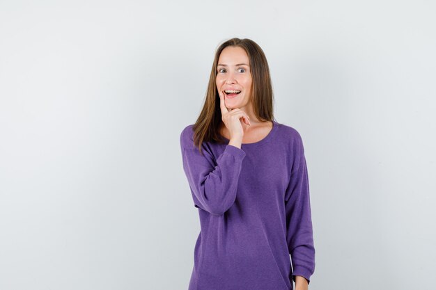Mujer joven en camisa violeta sosteniendo el dedo índice cerca de la boca y mirando feliz, vista frontal.