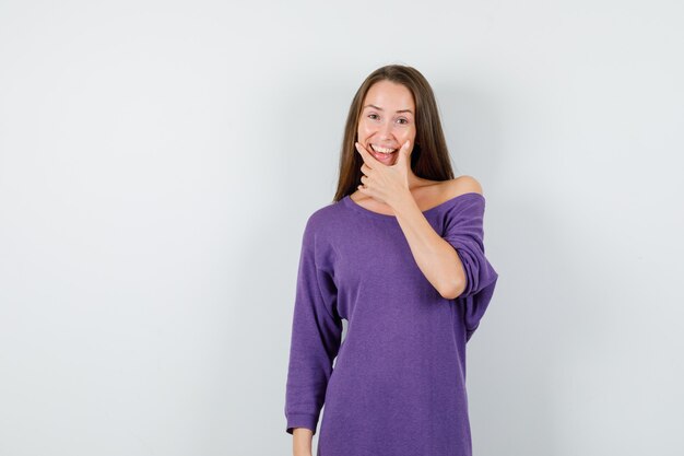 Mujer joven en camisa violeta sosteniendo la mano sobre la barbilla y mirando optimista, vista frontal.