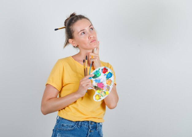 Mujer joven en camiseta amarilla, jeans mirando hacia arriba y sosteniendo herramientas de pintura