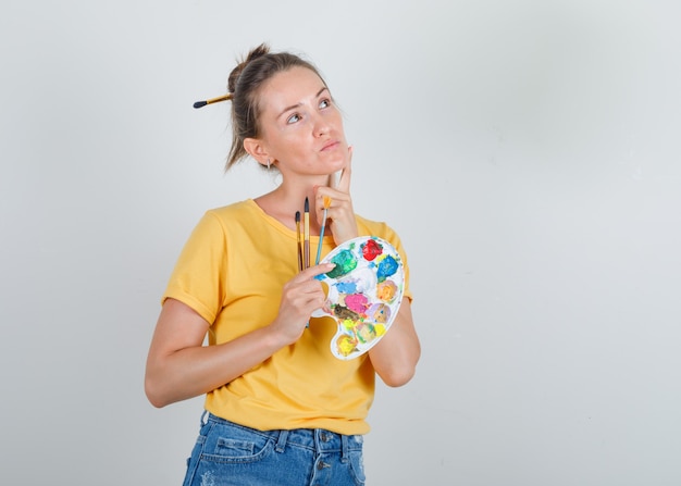 Mujer joven en camiseta amarilla, jeans mirando hacia arriba y sosteniendo herramientas de pintura