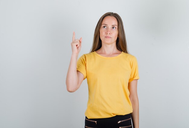 Mujer joven en camiseta amarilla, pantalones apuntando con el dedo hacia arriba y mirando pensativo