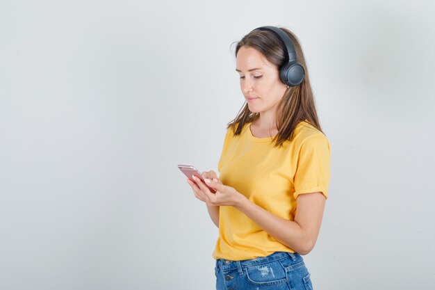 Mujer joven en camiseta amarilla, pantalones cortos escuchando música con auriculares
