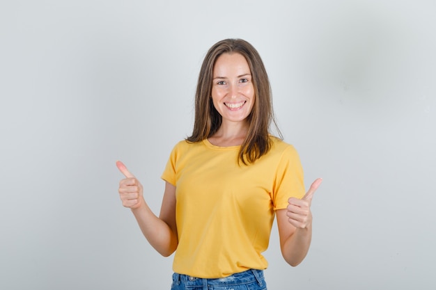 Mujer joven en camiseta amarilla, pantalones cortos mostrando los pulgares hacia arriba y mirando feliz