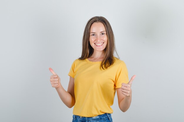 Mujer joven en camiseta amarilla, pantalones cortos mostrando los pulgares hacia arriba y mirando feliz