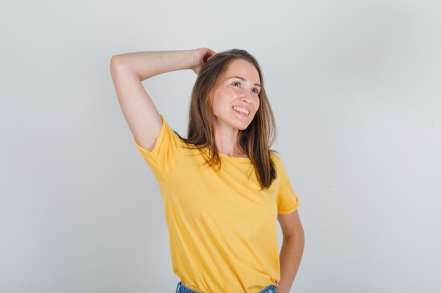 Mujer joven en camiseta amarilla, pantalones cortos pensando con las manos en la cabeza y mirando alegre