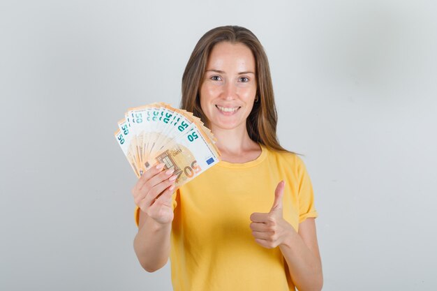 Mujer joven en camiseta amarilla, sosteniendo billetes en euros con el pulgar hacia arriba y mirando feliz