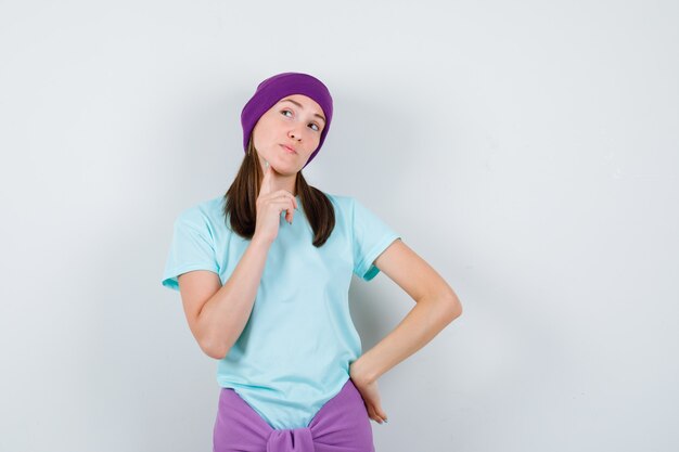 Mujer joven con camiseta azul, gorro morado con el dedo índice en la papada, poniendo una mano en la cadera, pensando en algo y mirando pensativo, vista frontal.