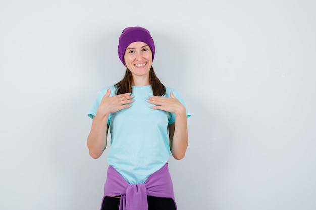 Mujer joven en camiseta azul, gorro morado con las manos sobre el pecho y mirando alegre, vista frontal.