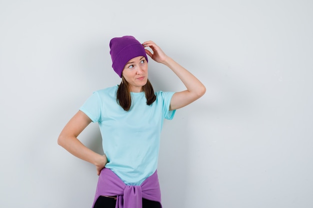 Mujer joven en camiseta azul, gorro morado rascándose la cabeza, con la mano en la cadera, pensando en algo y mirando pensativo, vista frontal.