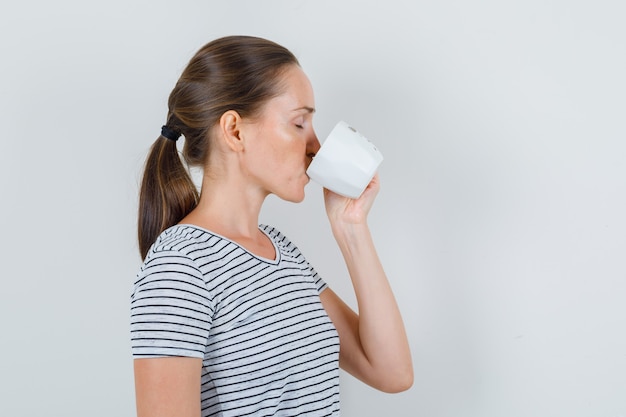 Mujer joven en camiseta bebiendo té y cerrando los ojos.