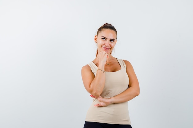 Mujer joven en camiseta beige de pie en pose de pensamiento y mirando bonita, vista frontal.