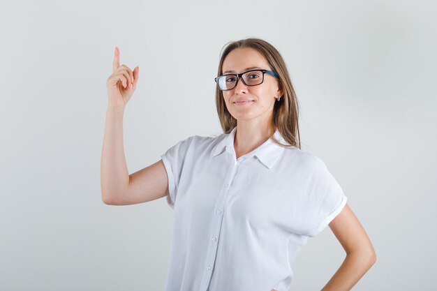 Mujer joven en camiseta blanca apuntando hacia arriba con el dedo y mirando alegre