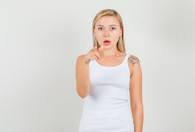 Mujer joven en camiseta blanca apuntando con el dedo a la cámara y mirando confundido