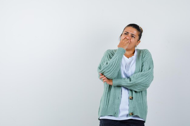 Mujer joven en camiseta blanca y cardigan verde menta de pie en pose de pensamiento y mirando pensativo