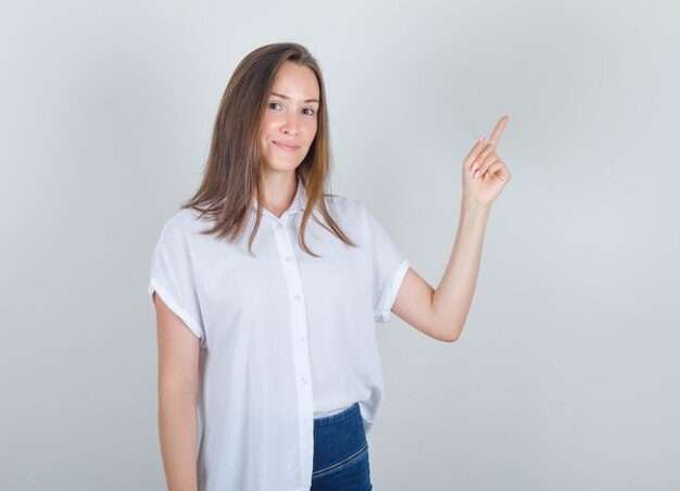 Mujer joven en camiseta blanca, jeans apuntando con el dedo y mirando contento