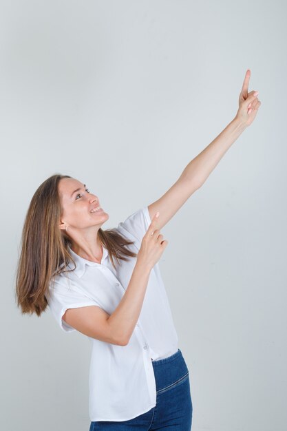 Mujer joven en camiseta blanca, jeans apuntando con los dedos y mirando alegre