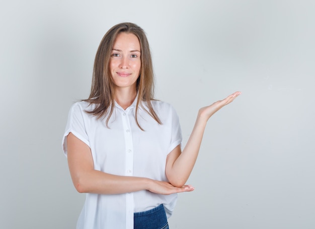 Mujer joven en camiseta blanca, jeans dando la bienvenida o mostrando algo y mirando contento