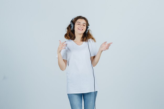 Mujer joven en camiseta blanca, jeans disfrutando de la música con auriculares y mirando feliz, vista frontal.