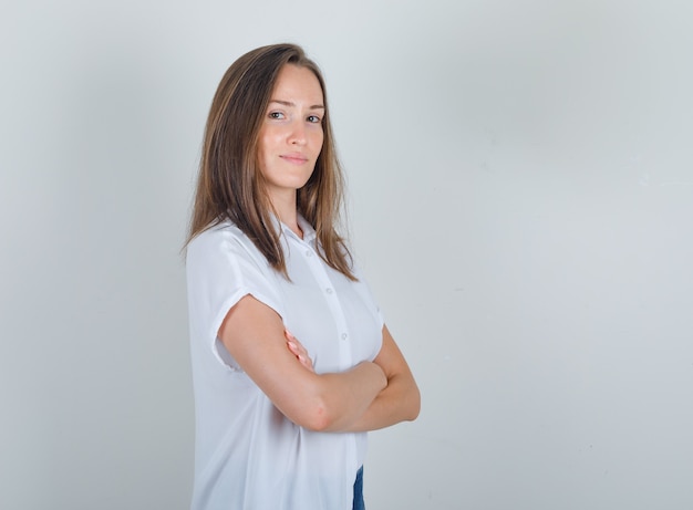 Mujer joven en camiseta blanca, jeans de pie con los brazos cruzados y mirando alegre.