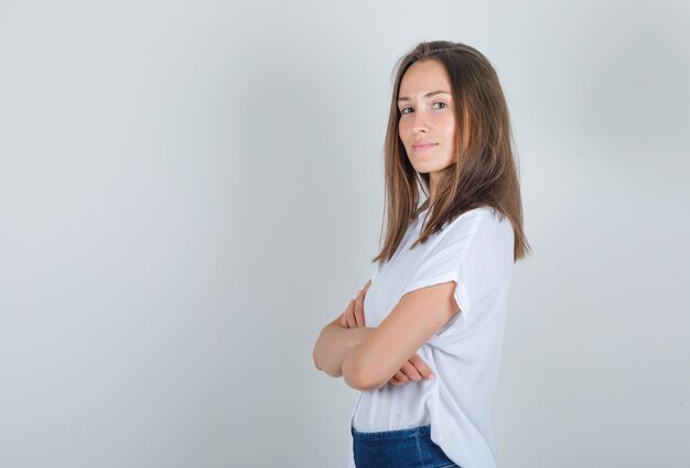 Mujer joven en camiseta blanca, jeans de pie con los brazos cruzados y mirando alegre.