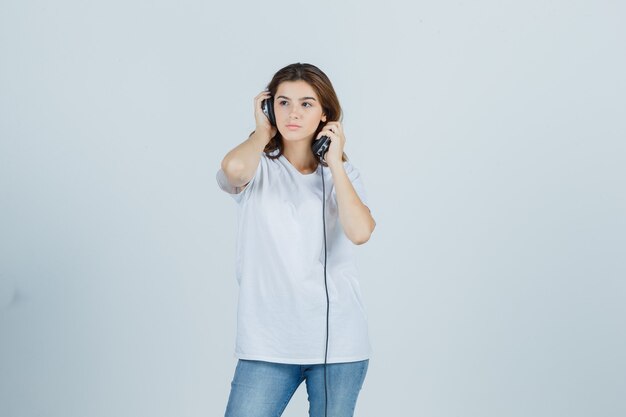 Mujer joven en camiseta blanca, jeans quitándose los auriculares y mirando pensativo, vista frontal.