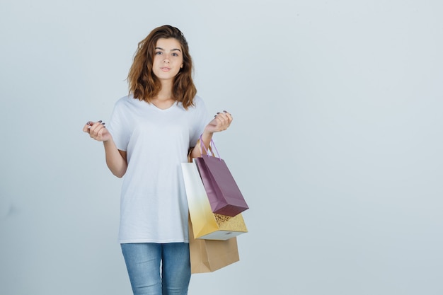 Mujer joven en camiseta blanca, jeans sosteniendo bolsas de papel y mirando encantador, vista frontal.