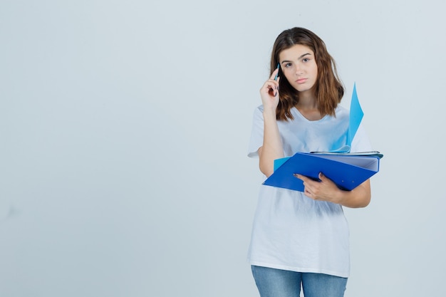 Mujer joven en camiseta blanca, jeans sosteniendo carpetas y bolígrafo y mirando pensativo, vista frontal.