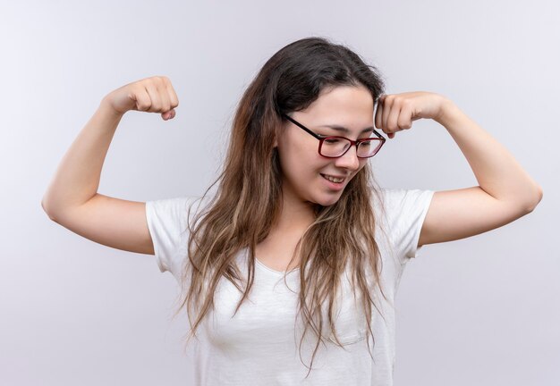 Mujer joven en camiseta blanca levantando el puño mostrando bíceps posando como un ganador positivo y feliz de pie sobre la pared blanca