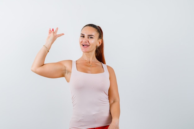 Mujer joven en camiseta blanca sin mangas, pantalones mostrando el signo de la victoria y mirando positivo, vista frontal.