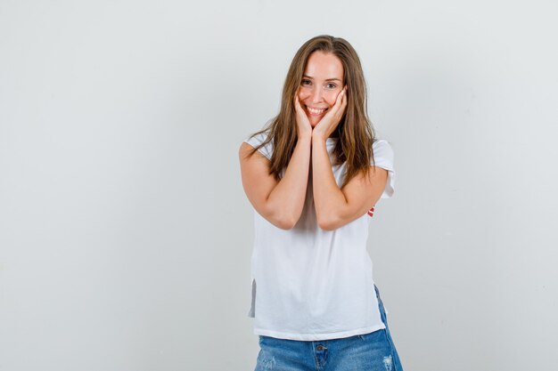 Mujer joven en camiseta blanca, pantalones cortos cogidos de la mano en las mejillas y sonriendo