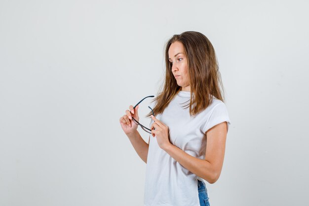 Mujer joven en camiseta blanca, pantalones cortos con gafas mientras piensa.