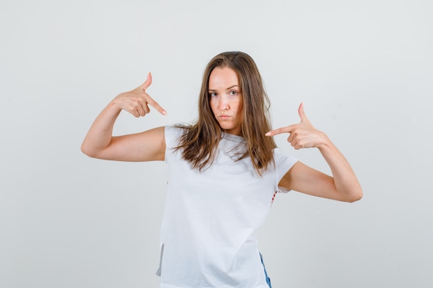 Mujer joven en camiseta blanca, pantalones cortos mostrándose con gesto de pistola y mirando confiada