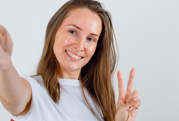 Mujer joven en camiseta blanca, pantalones cortos posando mientras muestra el signo de la victoria y parece feliz