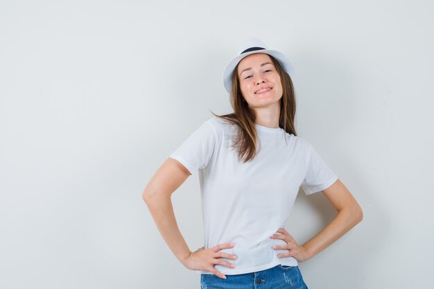 Mujer joven en camiseta blanca, pantalones cortos, sombrero posando mientras está de pie y parece seguro.