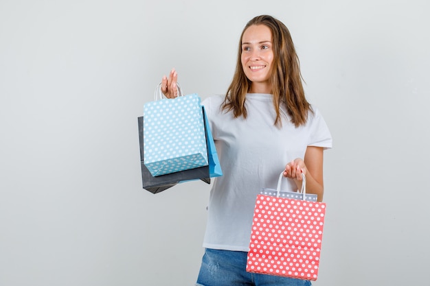 Mujer joven en camiseta blanca, pantalones cortos sosteniendo bolsas de papel mientras mira hacia otro lado y busca alegre