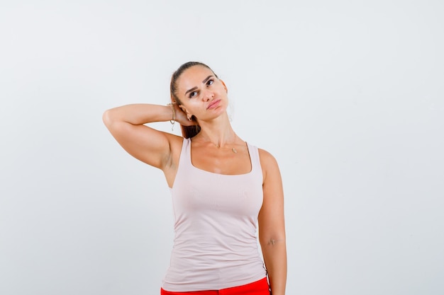 Foto gratuita mujer joven en camiseta blanca, pantalones sosteniendo la mano detrás de la cabeza y mirando pensativo, vista frontal.